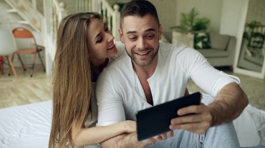 Tablet bilgisayar holding ve velilere evde yatakta oturmuş sohbet görüntülü sohbet sahip genç tatlı ve sevgi dolu Çift