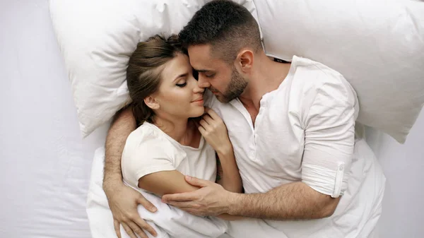 Junges schönes und liebevolles Paar küsst und umarmt sich im Bett, während es morgens aufwacht. Ansicht von oben attraktiver Mann küsst und spricht seine lächelnde Frau — Stockfoto