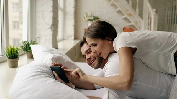 Jeune homme attrayant à l'aide d'un ordinateur tablette couché dans le lit tandis que sa petite amie venir et le câliner dans la chambre le matin — Photo