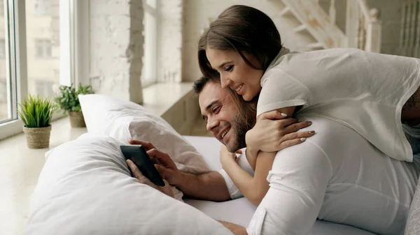 Young attractive man using tablet computer lying in bed while his girlfriend come and hug him in bedroom at the morning — Stock Photo, Image