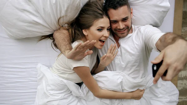 Fiatal szép és szerető pár miután online vidoe hívja smartphone kamera, miközben a reggel az ágyban fekve — Stock Fotó