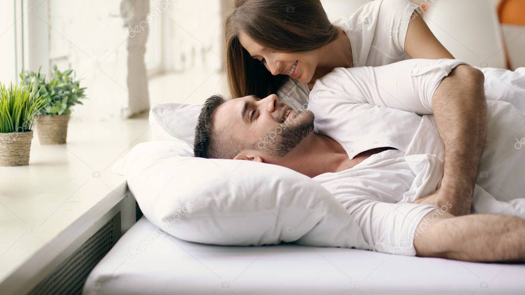 Young beautiful and loving couple wake up at the morning. Attractive woman kiss and hug his husband in bed