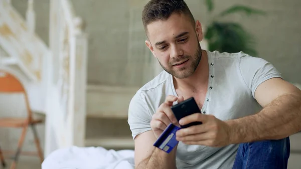 Vonzó fiatal férfi a smartphone és a hitelkártya-vásárlás az interneten üljön ágyban otthon — Stock Fotó