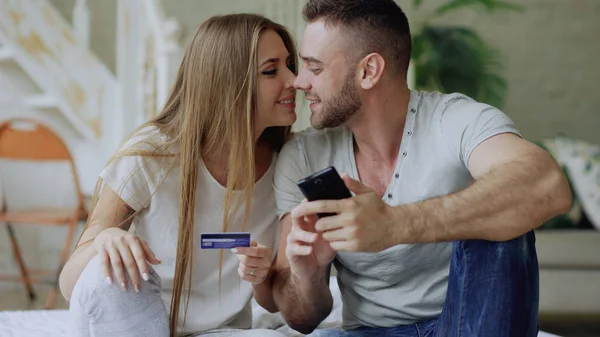 Atractiva pareja joven con teléfono inteligente y tarjetas de crédito de compras en Internet sentarse en la cama en casa — Foto de Stock