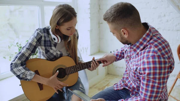 Молода щаслива і любляча пара вчиться грати на акустичній гітарі за допомогою планшетного комп'ютера і розважатися, сидячи за столом вдома — стокове фото
