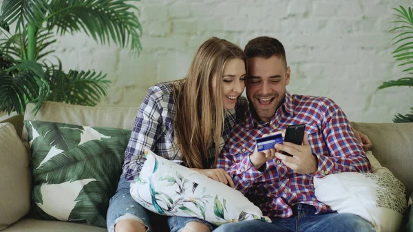 Pohledný mladý pár s smartphone a kreditní karty, nákup na internetu sedět na gauči v obývacím pokoji doma — Stock fotografie