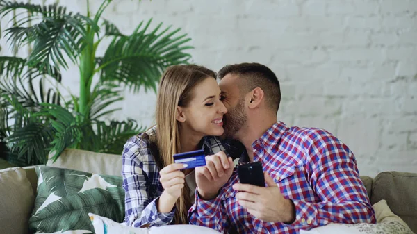 Attraktiva unga par med smartphone och kreditkort shopping på internet sitter på soffan i vardagsrummet hemma — Stockfoto