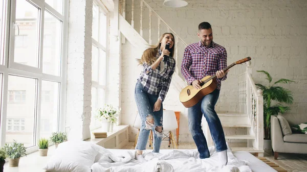 Vicces boldog és szerető pár tánc ágyon tv vezérlő énekelni és gitározni. Férfi és nő szórakozni nyaralás otthon — Stock Fotó