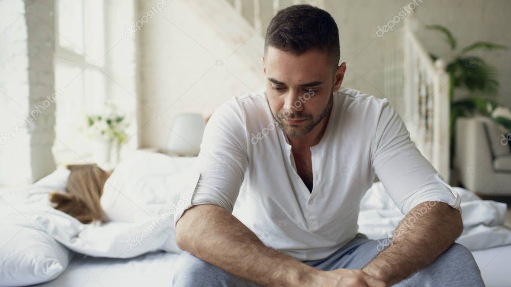 Upset young man sitting in bed suffer of problems while his girlfriend sleep in bedroom