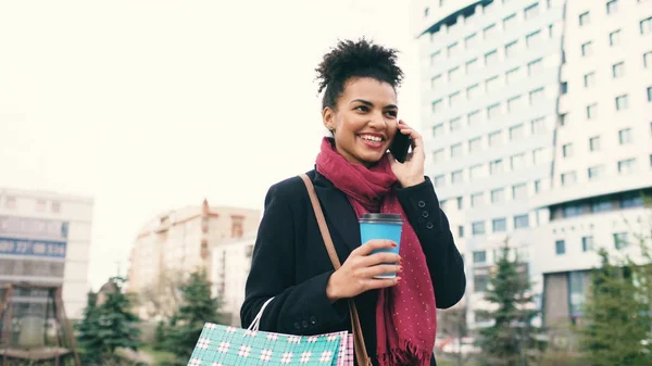 Привлекательная деловая женщина смешанной расы разговаривает со смартфоном и пьет кофе прогулки по городской улице с сумками — стоковое фото