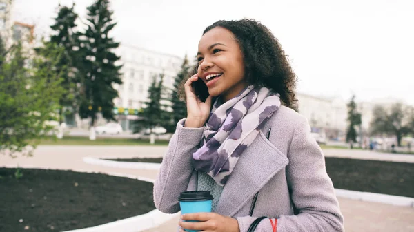 Привлекательная смешанная расовая девушка разговаривает на смартфоне и пьет кофе гуляет по городской улице с сумками. Молодая женщина идет после покупки в торговом центре продажи — стоковое фото
