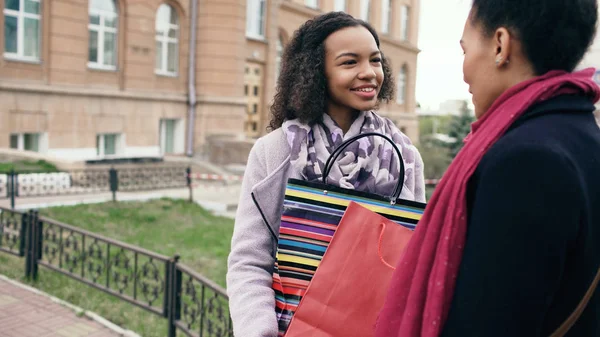 Две молодые африканские женщины делятся своими новыми покупками в пакетах друг с другом. Привлекательные девушки разговаривают и улыбаются после посещения торговых центров продаж — стоковое фото