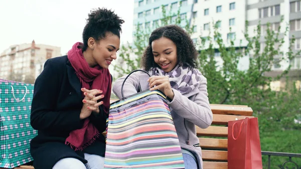 两个年轻非洲裔美国妇女彼此分享他们新的购买 shoppping 袋。迷人的女孩说话时坐在长椅上街 — 图库照片