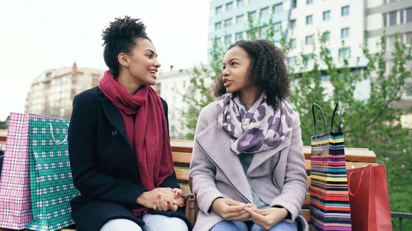 Две молодые африканские женщины делятся своими новыми покупками в пакетах друг с другом. Привлекательные девушки разговаривают, сидя на скамейке — стоковое фото