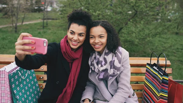 Dwa cute african american kobieta biorąc selfie na smartphone z torby na zakupy i uśmiecha się. Dziewczyny, siedząc na ławce ulicy miłej zabawy po wizycie w centrum sprzedaży — Zdjęcie stockowe