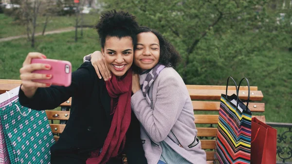 Две симпатичные африканские американки делают селфи на смартфоне с пакетами покупок и улыбаются. Девушки, сидящие на уличной скамейке, веселятся после посещения торговых центров — стоковое фото