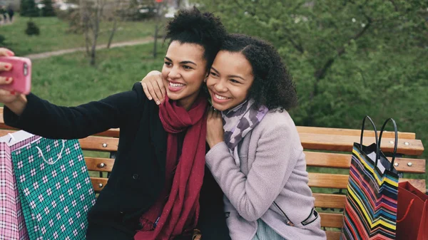 Dua wanita afrika lucu mengambil selfie di smartphone dengan tas belanja dan tersenyum. Girlfriends duduk di bangku jalan bersenang-senang setelah mengunjungi penjualan mal — Stok Foto