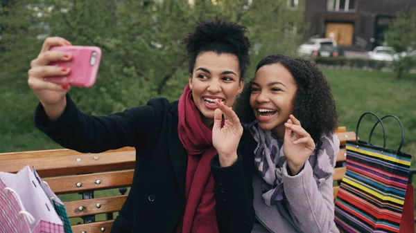 Dua wanita ras campuran ceria dengan tas belanja berbicara di panggilan video dengan smartpone. Gadis muda duduk di bangku memiliki chatting online dengan teman-teman — Stok Foto