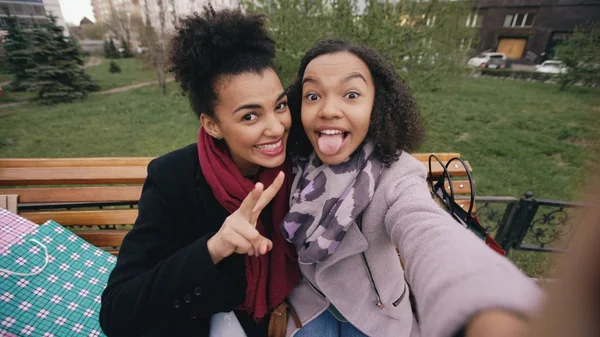 Dwa cute african american kobieta biorąc selfie na smartphone z torby na zakupy i uśmiecha się. Dziewczyny, siedząc na ławce ulicy miłej zabawy po wizycie w centrum sprzedaży — Zdjęcie stockowe