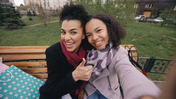 Две симпатичные африканские американки делают селфи на смартфоне с пакетами покупок и улыбаются. Девушки, сидящие на уличной скамейке, веселятся после посещения торговых центров — стоковое фото