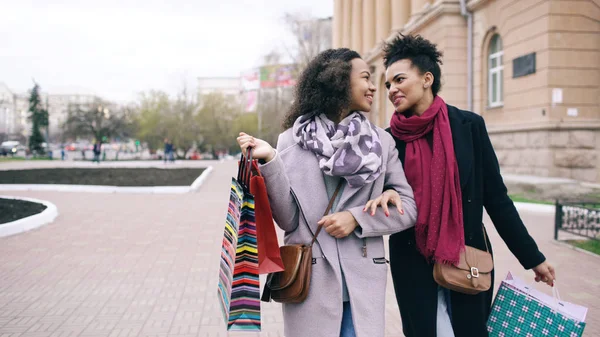 Две привлекательные женщины смешанной расы с сумками для покупок разговаривают и идут по улице. Подружки веселятся после продажи торгового центра — стоковое фото