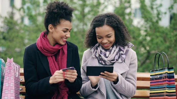 Twee aantrekkelijke gemengd ras vrouwen met papieren zakken zittend op de Bank en het gebruik van tablet pc en een creditcard voor online winkelen — Stockfoto