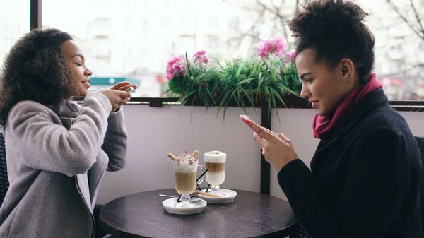 Dos atractivas amigas de raza mixta fotografiando tazas de café juntas usando la cámara del teléfono inteligente mientras están sentadas en la cafetería al aire libre —  Fotos de Stock