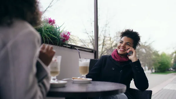 Dos mujeres de raza mixta sentadas a la mesa en el café de la calle usando un teléfono inteligente y hablando — Foto de Stock