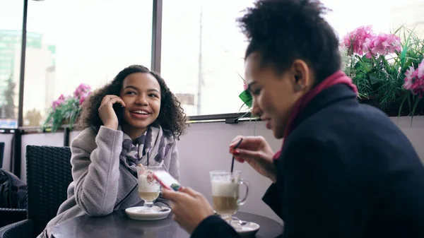 Dos mujeres de raza mixta sentadas a la mesa en el café de la calle usando un teléfono inteligente y hablando — Foto de Stock