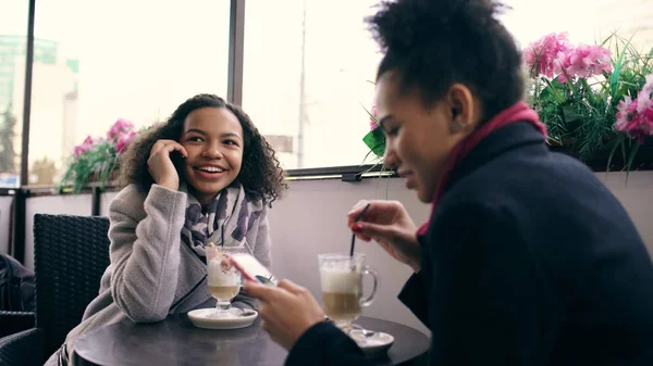 Dos mujeres de raza mixta sentadas a la mesa en el café de la calle usando un teléfono inteligente y hablando — Foto de Stock
