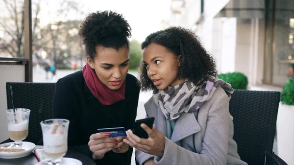 Two attractive mixed race women having online shopping with credit card and smartphone while talking and drinking coffee in street cafe — Stock Photo, Image