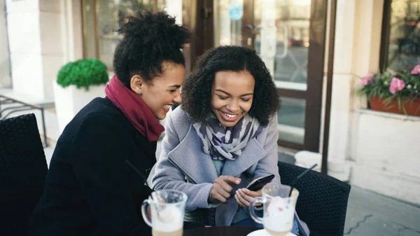 Twee aantrekkelijke gemengd ras-vriendinnen delen samen met behulp van smartphone in straat café in openlucht — Stockfoto