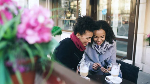 İki çekici karışık ırk kadın arkadaş birlikte sokak kafe mekanlarda smartphone kullanarak paylaşma — Stok fotoğraf
