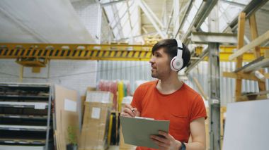 Happy young worker in industrial warehouse listening to music and dancing during work. Man in headphones have fun at workplace. clipart