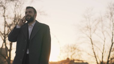 Genç iş adamı smartphone vasıl konuşuyor ve akşam saat içinde güneş ışığı arkadan aydınlatmalı yoIda fırsatlar yapma paltolu sakallı