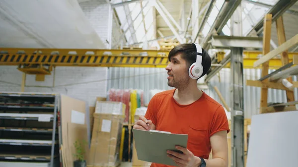 Happy young worker in industrial warehouse listening to music and dancing during work. Man in headphones have fun at workplace. — Stock Photo, Image