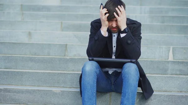 Молодой бизнесмен с ноутбуком испытывает стресс после телефонного звонка и сидит на лестнице на улице. Бизнесмен, имеющий проблемы на работе — стоковое фото
