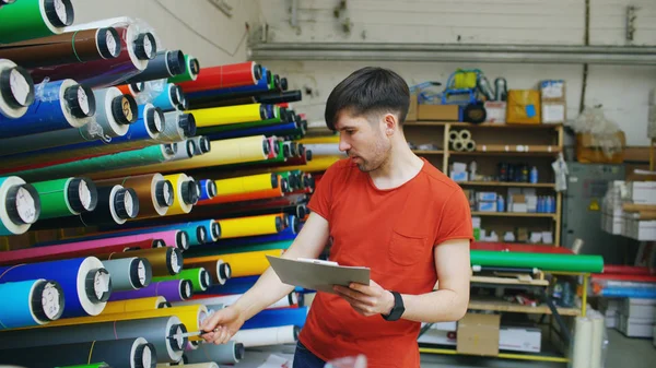 Young worker in warehouse with clipboard checking inventory. Man works in sales department of advertising materials — Stock Photo, Image