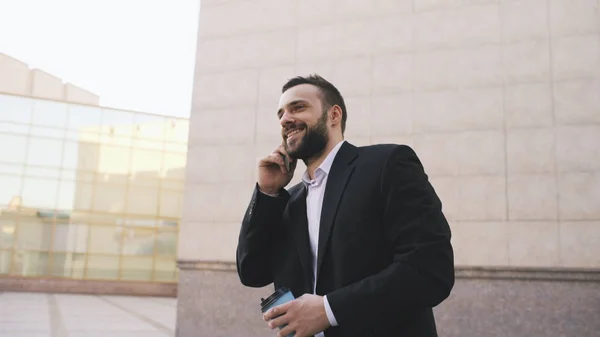 Joven hombre de negocios barbudo hablando por teléfono celular haciendo tratos con taza de café cerca de modernos edificios de oficinas — Foto de Stock