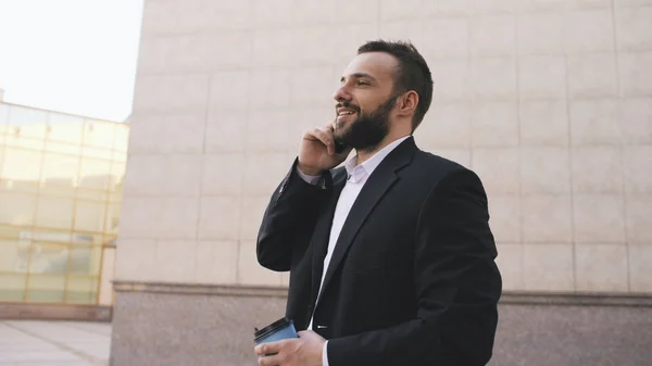 Fiatal szakállas üzleti ember foglalkozik csésze kávé közelében irodaépületek mobiltelefonon beszél — Stock Fotó
