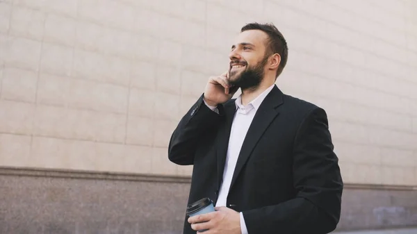 Joven hombre de negocios barbudo hablando por teléfono celular haciendo tratos con taza de café cerca de modernos edificios de oficinas — Foto de Stock