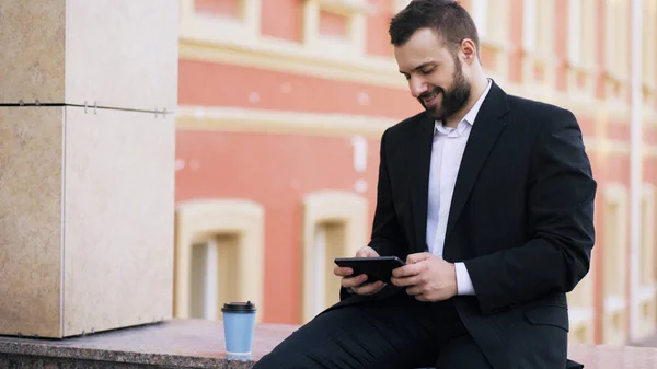 Fiatal szakállas üzletember olvasó Hírek-ra tabletta számítógép és ital kávé szünet a hivatali épület közelében — Stock Fotó