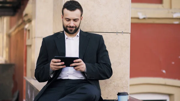 Jeune homme d'affaires barbu lire des nouvelles sur tablette et boire du café pendant la pause près de son immeuble de bureaux — Photo