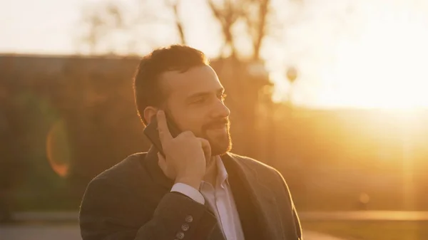 Closeup mladých vousatých obchodní muž v plášti mluvit na smartphone a dělání nabídky na ulici s podsvícením slunce ve večerních hodinách — Stock fotografie