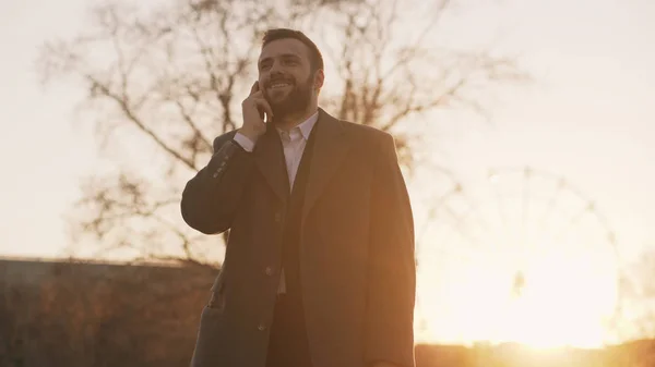 Giovane uomo d'affari barbuto in cappotto parlare allo smartphone e fare offerte su strada con la luce del sole retroilluminato in tempo serale — Foto Stock