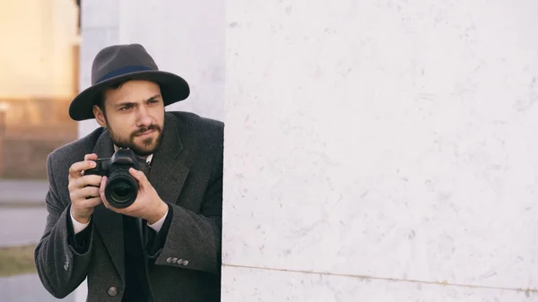 Jeune agent espion masculin portant chapeau et manteau photographiant des criminels et se cachant derrière le mur — Photo