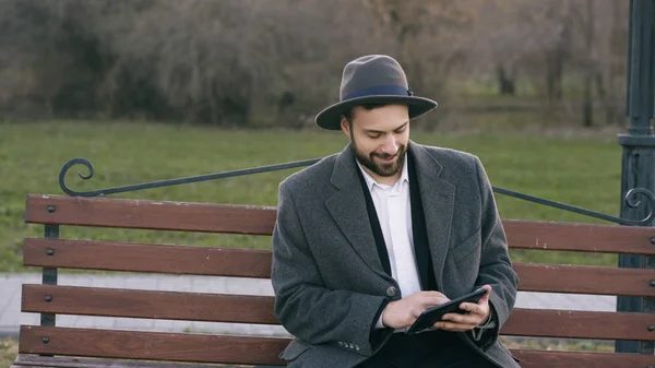Hipser twórczej działalności człowieka w kapelusz za pomocą komputera typu tablet pc i siedzi na ławce ulicy miasta, w parku podczas przerwy — Zdjęcie stockowe