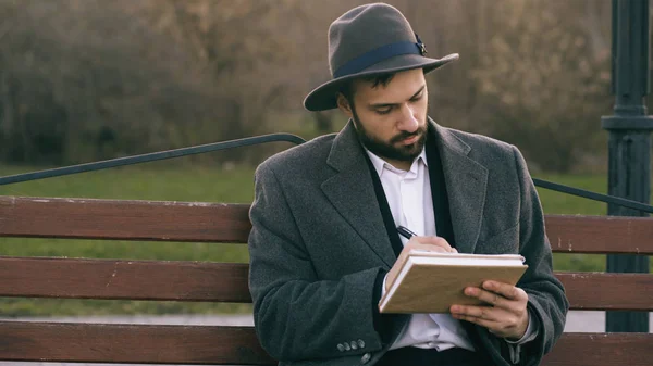 Hipser artiste créatif homme en chapeau et manteau dessin avec stylo et carnet de croquis assis sur le banc de rue de la ville au parc — Photo