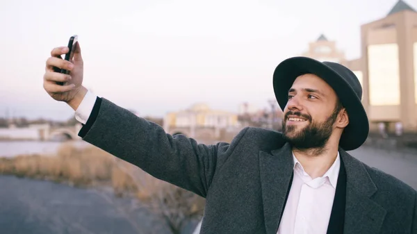 Mladí rádi turistické muž v ahat a kabát s úsměvem při pořizování selfie obrázek s mobilním telefonem na město u řeky — Stock fotografie