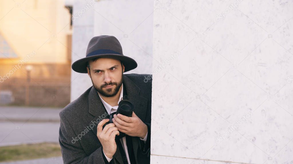Young male spy agent wearing hat and coat photographing criminal people and hiding behind the wall